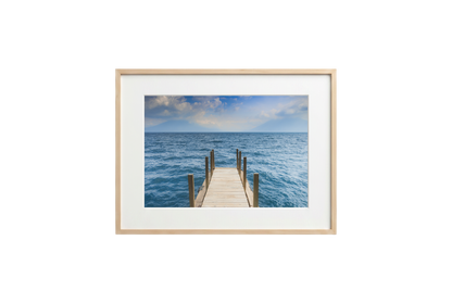 Framed photograph of a dock extending into the ocean on a white wall.