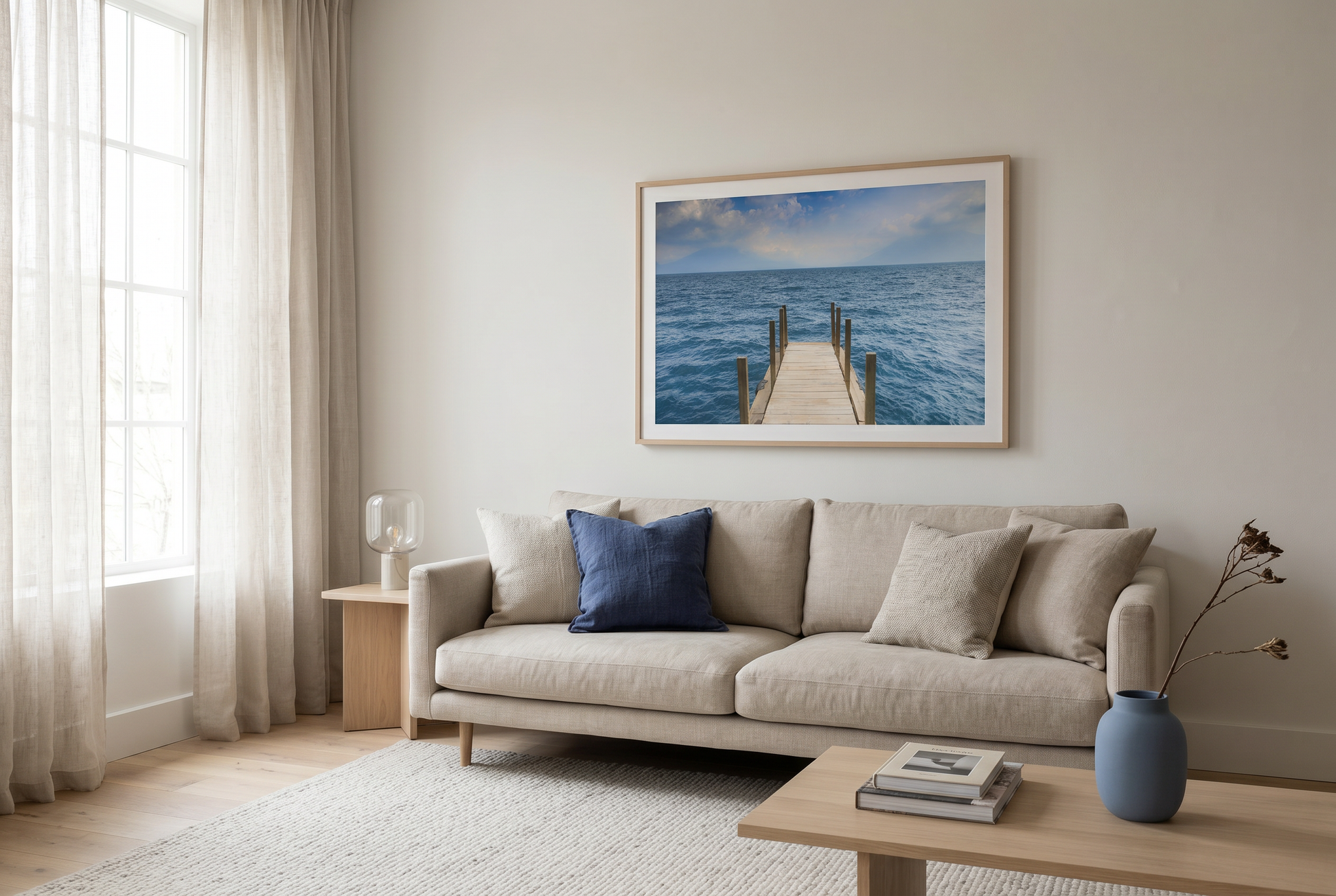 Living room with beige sofa, blue pillow, and framed photograph of a dock extending into the ocean on a white wall.picture on wall.