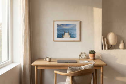 Home office with wooden desk, chair, and Framed photograph of a dock extending into the ocean on a white wall..