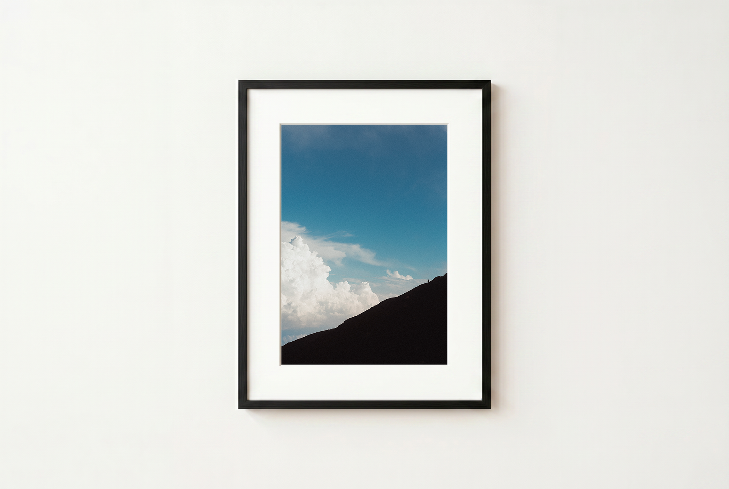 Framed photograph of a mountain landscape with blue sky and clouds on a white wall.