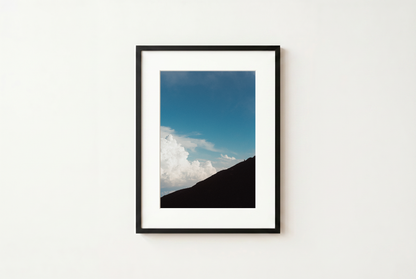 Framed photograph of a mountain landscape with blue sky and clouds on a white wall.