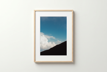Framed photograph of a mountain landscape with blue sky and clouds on a white wall.
