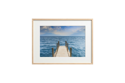 Framed photograph of a dock extending into the ocean on a white wall.