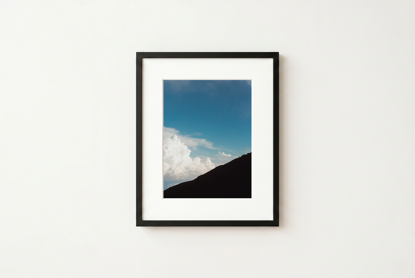 Framed photograph of a mountain with a blue sky and clouds on a white wall.