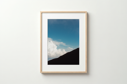Framed photograph of a mountain and blue sky with clouds on a white wall.