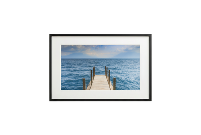 Framed photograph of a dock extending into the ocean on a white wall.