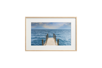Framed photograph of a dock extending into the ocean on a white wall.