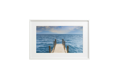 Framed photograph of a dock extending into the ocean on a white wall.