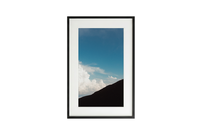 Framed picture of a mountain landscape with blue sky and clouds.