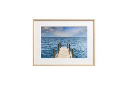 Framed photograph of a dock extending into the ocean on a white wall.