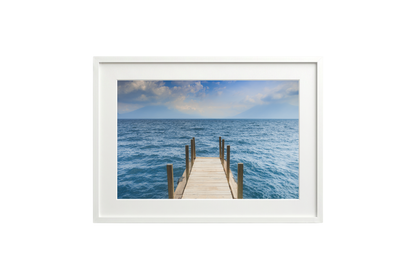 Framed photograph of a dock extending into the ocean on a white wall.