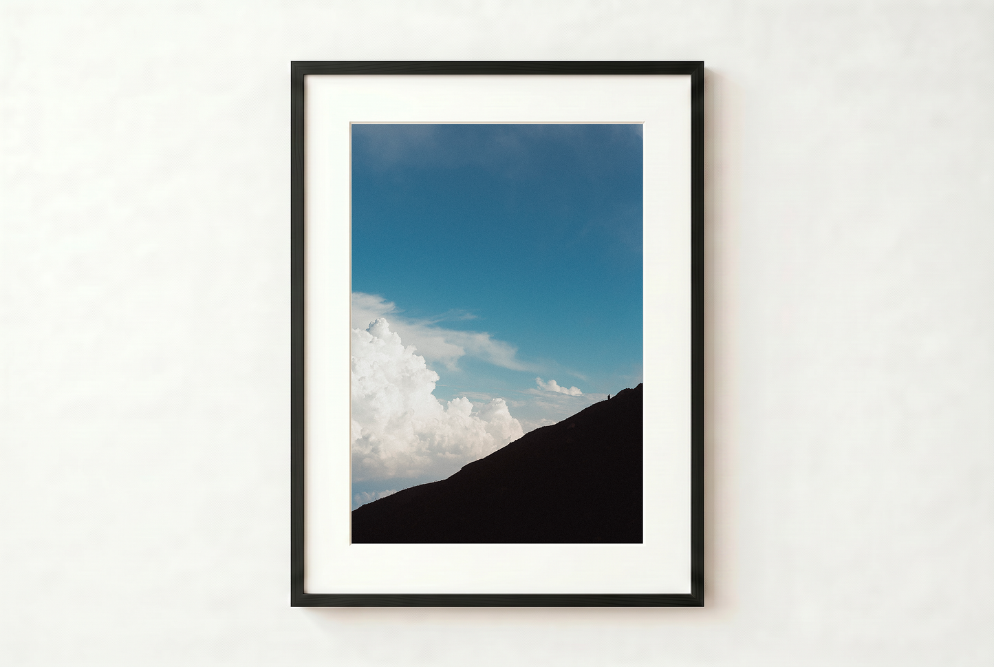Framed photograph of a mountain and blue sky with clouds on a white wall.