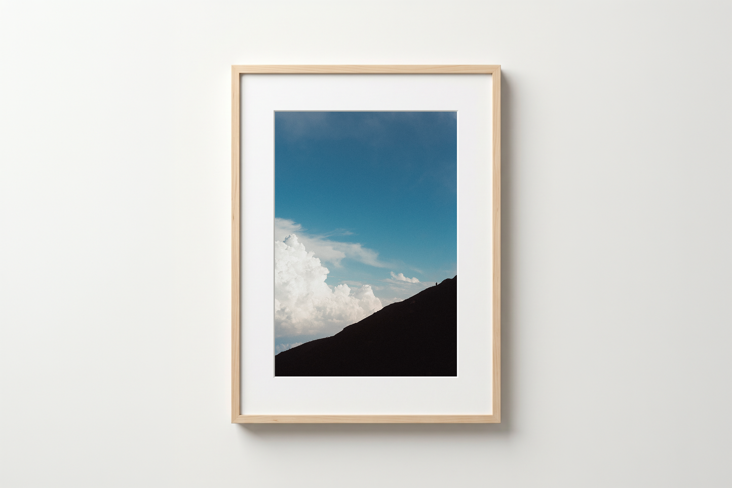 Framed photograph of a mountain landscape with blue sky and clouds on a white wall.