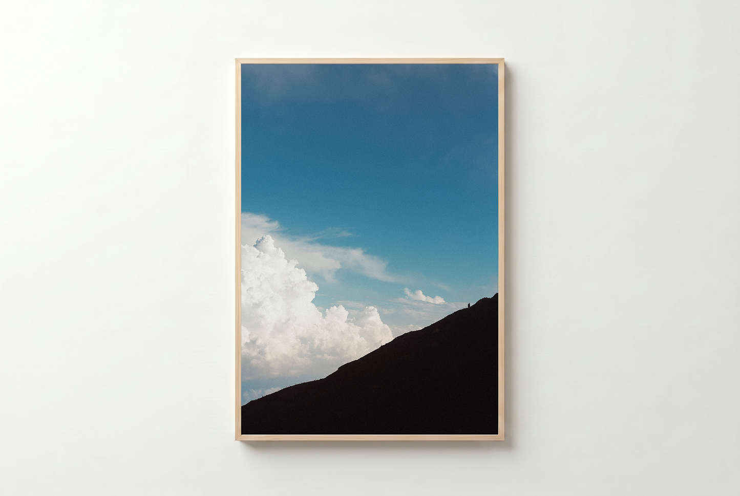 Framed artwork of a mountain silhouette against a blue sky with clouds on a white wall.