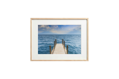 Framed photograph of a dock extending into the ocean on a white wall.