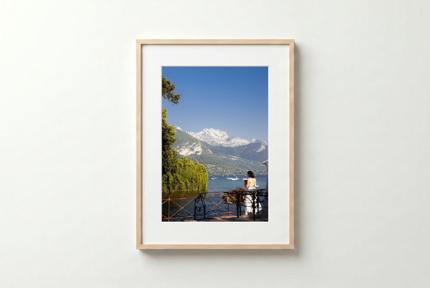 Framed photograph of a person sitting by a lake with mountains in the background on a white wall.