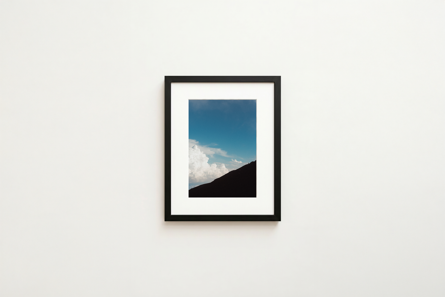 Framed artwork of a mountain landscape with blue sky and clouds on a white wall.