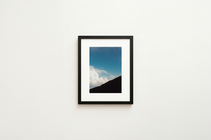 Framed artwork of a mountain landscape with blue sky and clouds on a white wall.