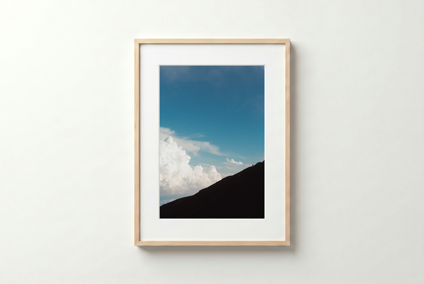 Framed photograph of a mountain landscape with blue sky and clouds on a white wall.