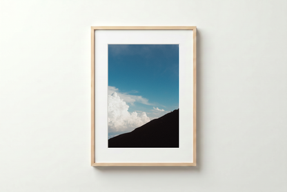 Framed photograph of a mountain landscape with blue sky and clouds on a white wall.