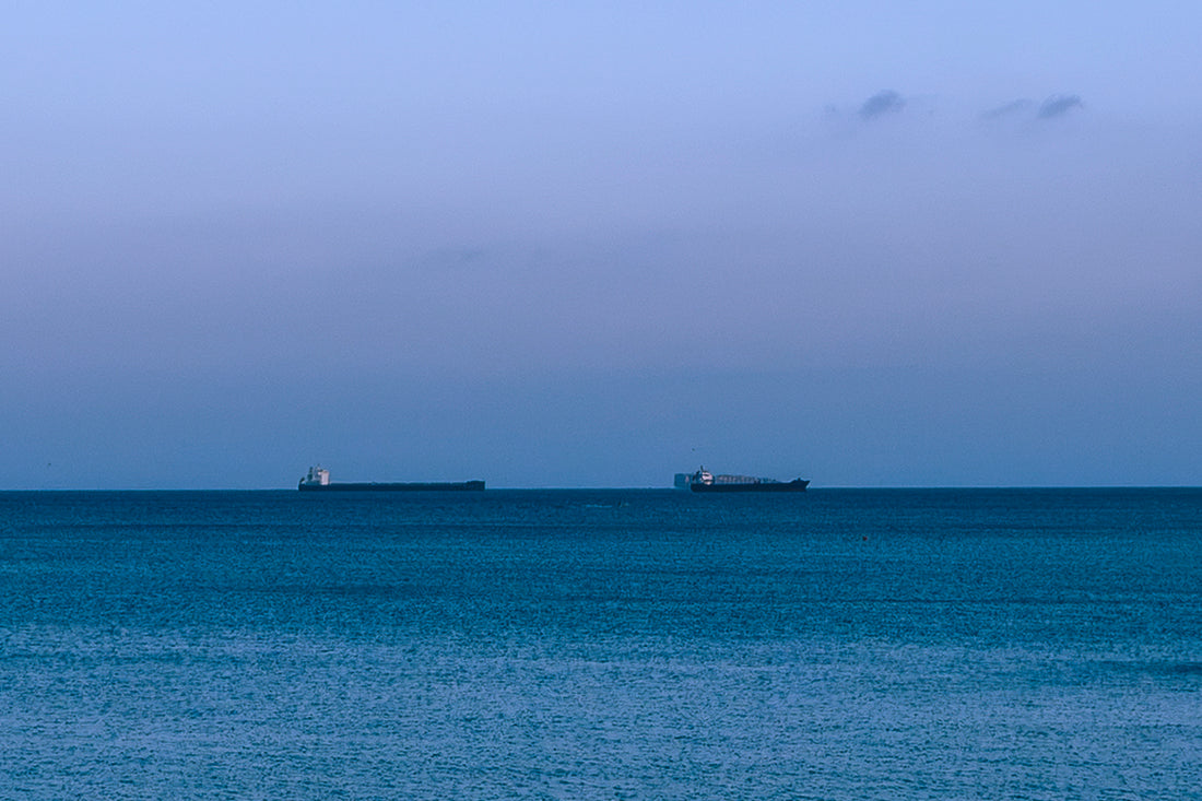 
A wide seascape with calm blue water under a pale blue sky, with two small cargo ships visible on the horizon.