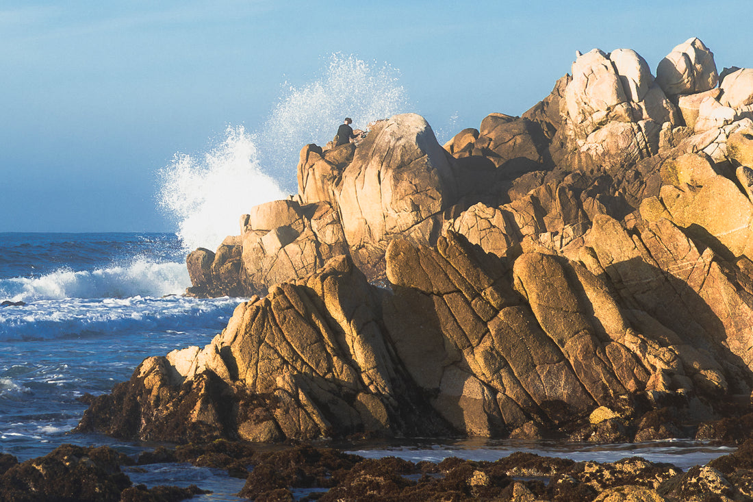 
A dramatic coastal scene with large tan and brown rock formations rising from the ocean, white sea spray at the top of the cliffs, blue water, and a pale blue sky. A small person is visible sitting on the highest rock formation.