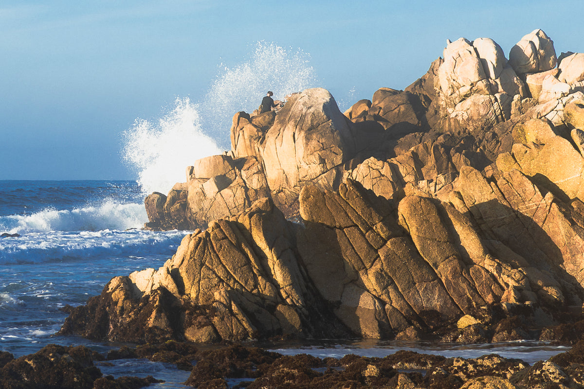 
A dramatic coastal scene with large tan and brown rock formations rising from the ocean, white sea spray at the top of the cliffs, blue water, and a pale blue sky. A small person is visible sitting on the highest rock formation.