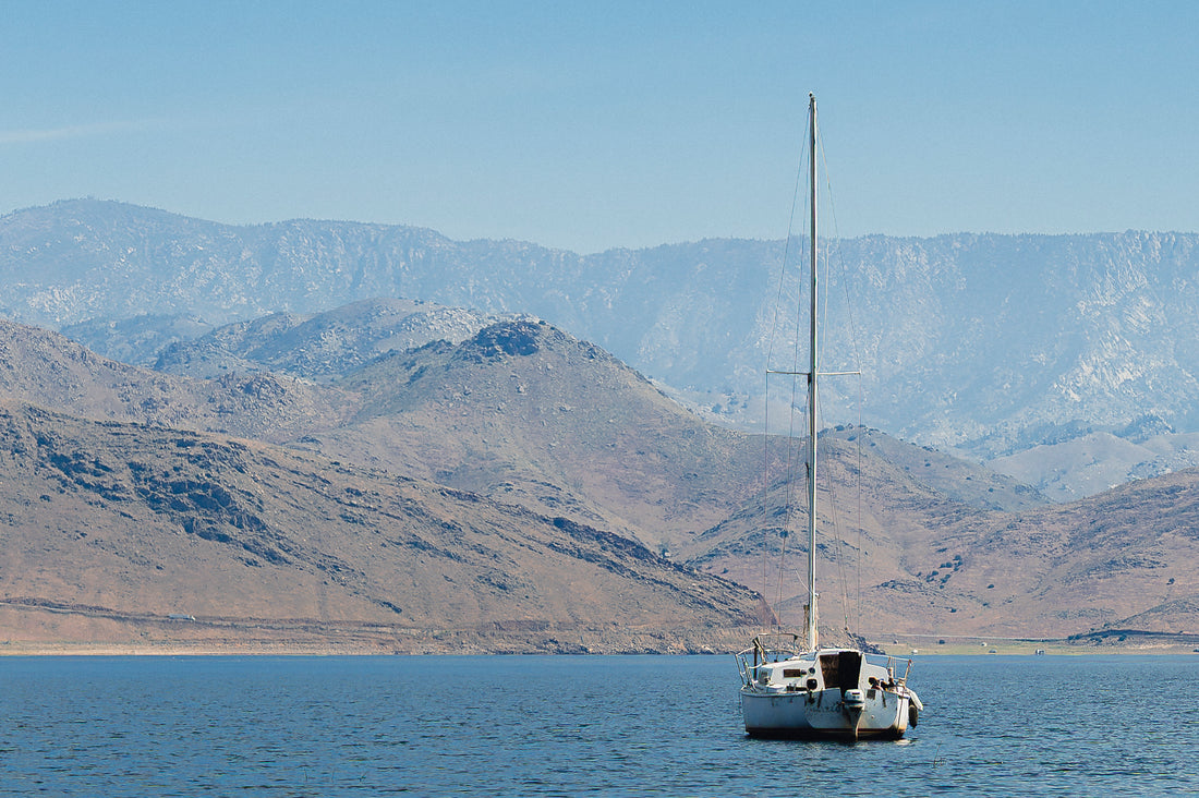 
A white sailboat with its mast raised is anchored on calm blue water, with arid brown mountains and hazy blue peaks in the background under a clear sky.
