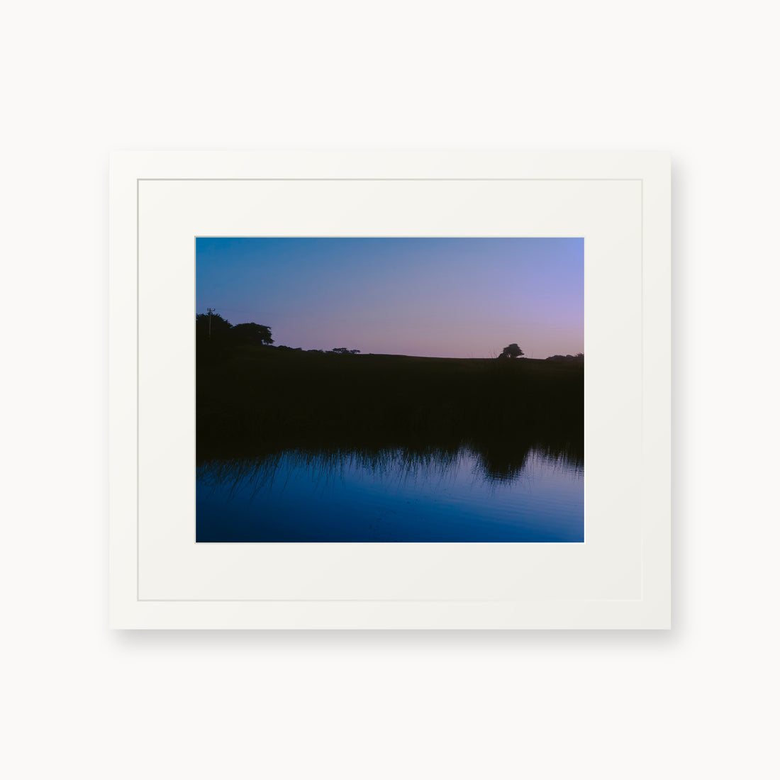 
What Lays Beneath — 11x14, white frame. Dark trees and a twilight shoreline reflected across blue water.
