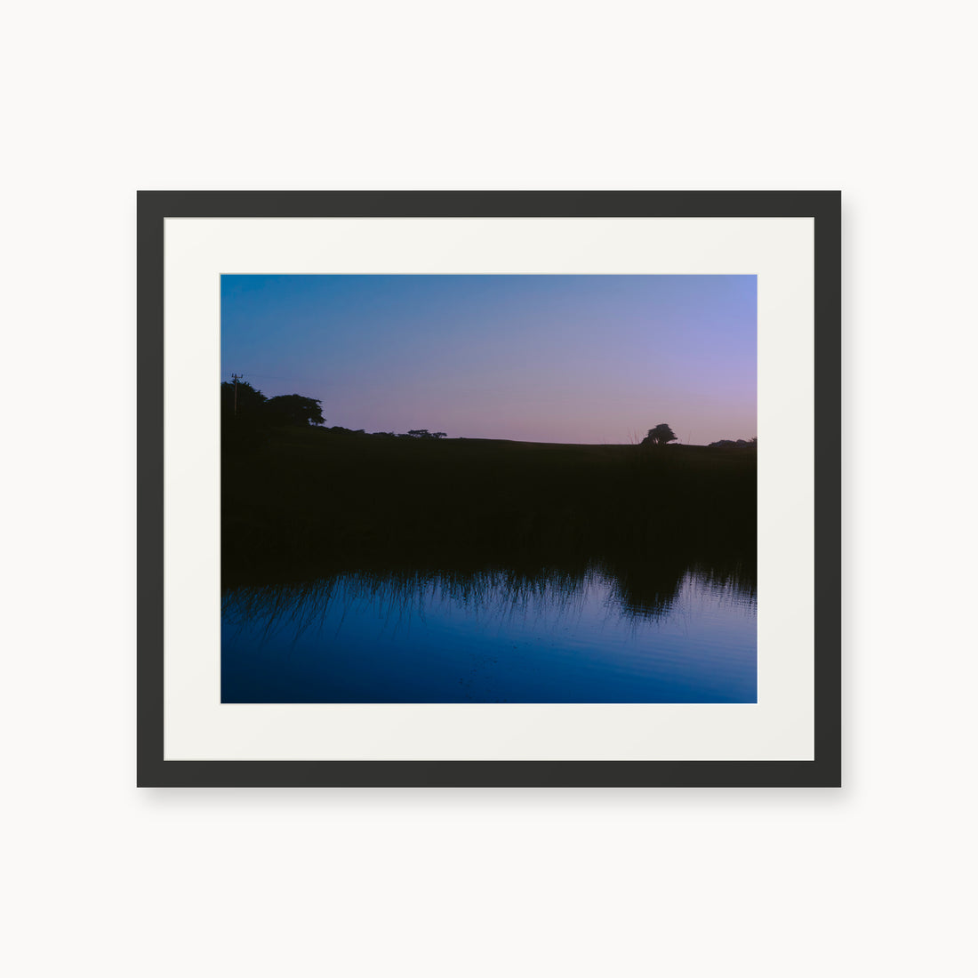
What Lays Beneath — 16x20, black frame. Dark trees and a twilight shoreline reflected across blue water.