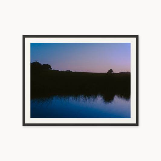 
What Lays Beneath — 30x40, black frame. Dark trees and a twilight shoreline reflected across blue water.