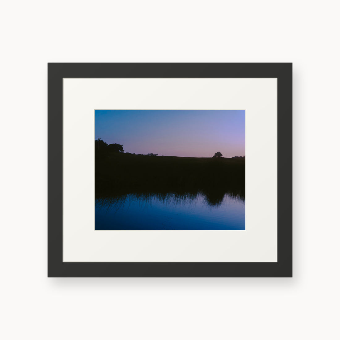 
What Lays Beneath — 8x10, black frame. Dark trees and a twilight shoreline reflected across blue water.