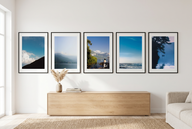 Five framed blue toned calming artworks on a wall above a wooden cabinet in a modern living room with a large window and sofa.