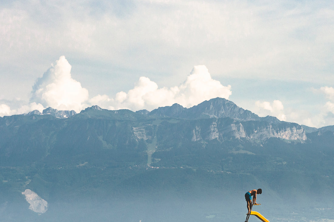 
Person standing on a diving board with mountains in the background of a serene beach scene.